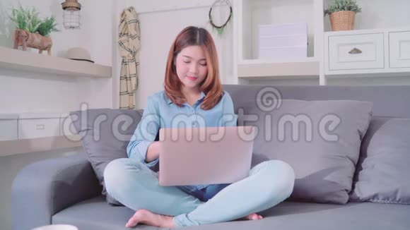 迷人的年轻微笑亚洲妇女使用电脑或笔记本电脑当躺在沙发上放松在客厅在家里视频的预览图