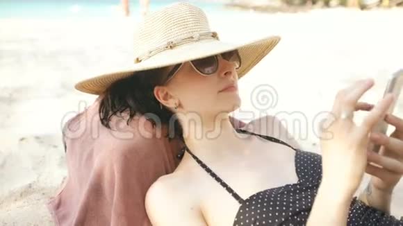 戴着帽子戴着太阳镜的年轻女人用智能手机躺在海滩上视频的预览图