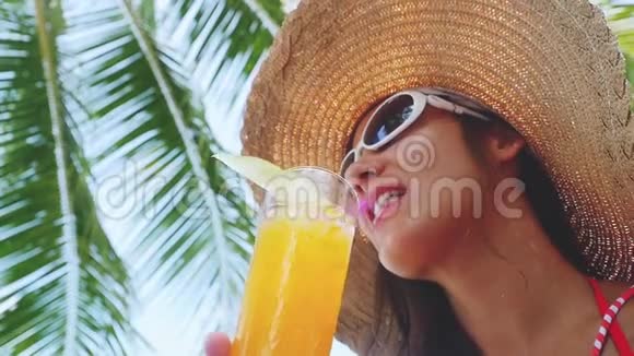 美丽的年轻女子戴着太阳镜和帽子在海滩上喝热带鸡尾酒在阳光明媚的天气里慢慢地享受着阳光视频的预览图