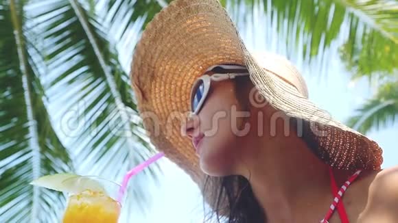 美丽的黑发女人戴着太阳镜和帽子带着热带鸡尾酒在海滩上享受阳光明媚的天气在缓慢的天气里视频的预览图
