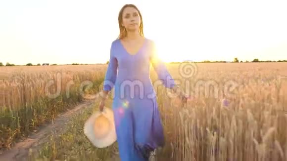 美丽的女人穿着蓝色的连衣裙和帽子在日落时穿过麦田自由概念日落时的麦田视频的预览图