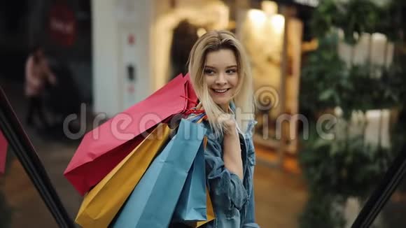 穿着牛仔裤夹克的金发女郎微笑着走下购物中心的移动楼梯视频的预览图