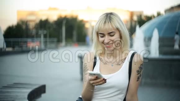 年轻的金发女郎坐在长凳上看着相机和微笑的肖像户外视频的预览图
