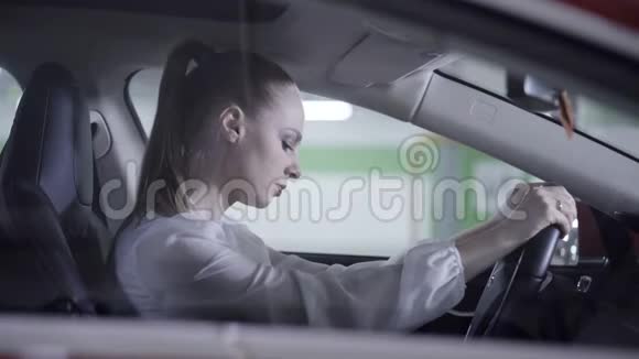 白人女性穿着白衬衫坐在车里拿着方向盘在车库旁边视频的预览图