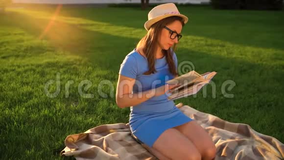 戴眼镜的女孩在日落时坐在公园的毯子上看书视频的预览图