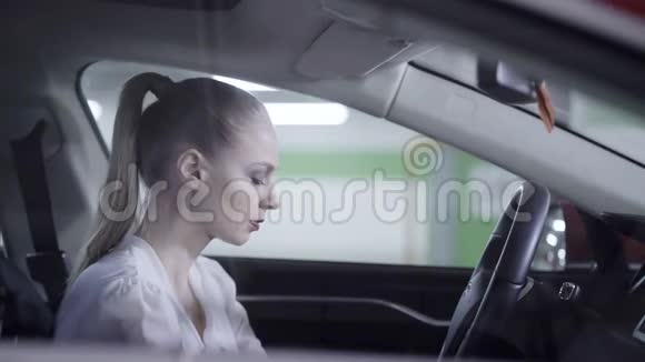 年轻的女性穿着白色衬衫坐在车里拿着方向盘在车库旁边视频的预览图