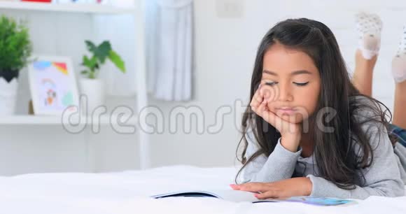 美丽的亚洲年轻的黑发女人在家里的床上看书微笑着面对视频的预览图