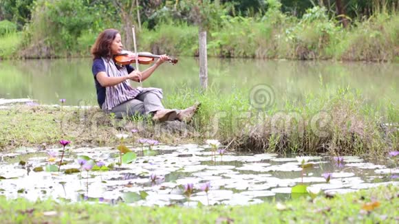 一位留着黑发牛仔裤和围巾的亚洲女人在湖边拉小提琴视频的预览图