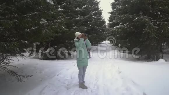 美丽的年轻女子穿着冬天的衣服手套和羊毛帽用针织相机拍摄雪林视频的预览图