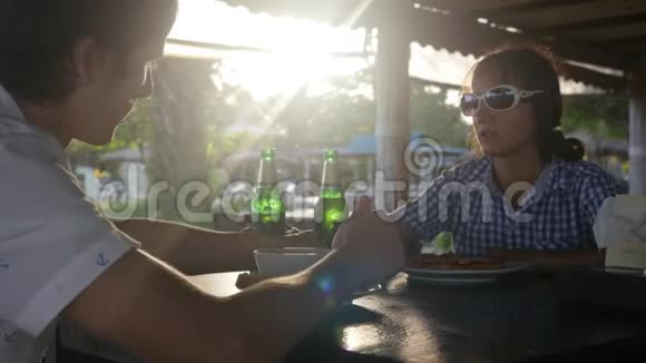 快乐的年轻夫妇吃着饭喝着啤酒在阳光明媚的时候在咖啡馆里用镜头闪烁的效果拍打着瓶子视频的预览图