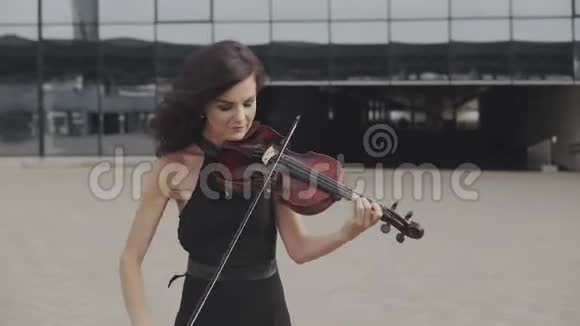 优雅的女人穿着裙子在玻璃大厦附近拉小提琴城市艺术概念视频的预览图