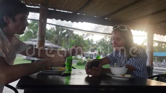 快乐的年轻夫妇在户外咖啡馆里吃着饭喝着啤酒女孩在夏日的智能手机上展示了一些东西视频的预览图