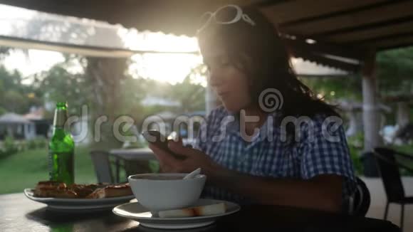 在日落时分喝啤酒的年轻女子在户外咖啡馆里吃晚饭并用手机发送信息视频的预览图