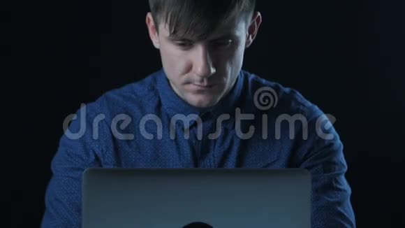 一个穿着蓝色衬衫的男人在黑暗房间里的笔记本电脑上工作的肖像视频的预览图