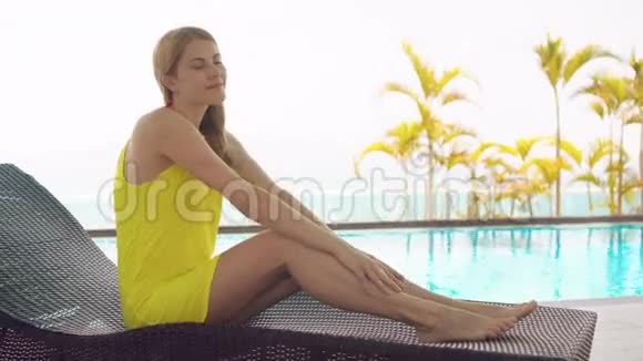 一位穿着黄色衣服躺在绿松石水游泳池旁的日光浴池里的美丽梦幻的年轻女子视频的预览图