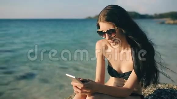 美丽的女性在度假期间在海滩日落时使用智能手机短信视频的预览图