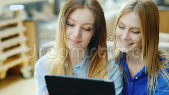 两个年轻的女朋友坐在咖啡馆里用平板电脑有兴趣看展示积极的情绪视频的预览图