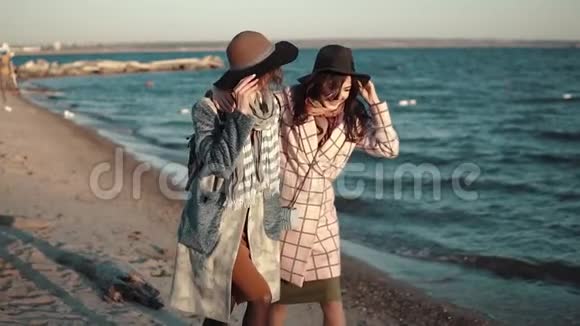两个女朋友高兴地跑来跑去在露天嬉戏日落时穿着秋装和帽子的年轻女子在海滩上视频的预览图