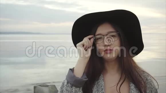 日落时分戴着黑帽子戴着眼镜眼睛在海边眨眨眼的年轻美女视频的预览图