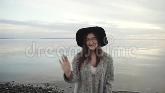 日落时分戴着黑帽子戴着眼镜的年轻漂亮女子在海边挥手致意视频的预览图