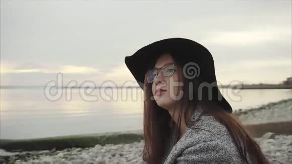 年轻漂亮的女人戴着黑色的帽子和眼镜坐在海边日落附近视频的预览图