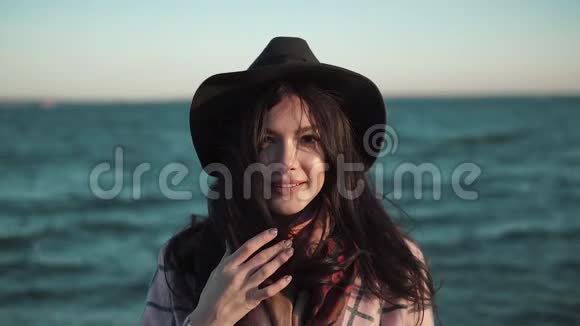 海滩上一个年轻女人的肖像戴着帽子和秋装的女孩微笑着在水边的摄像机上摆姿势慢慢视频的预览图