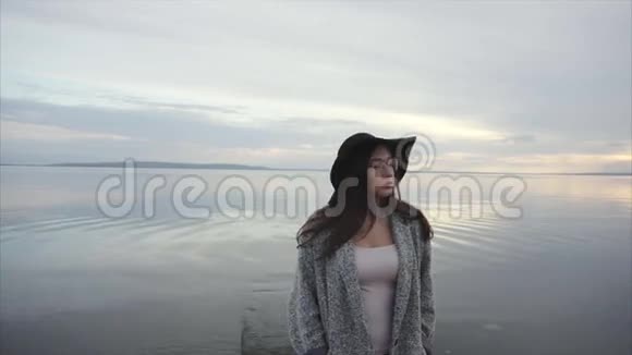 戴着黑色帽子和眼镜的年轻女子在日落时环视海岸视频的预览图