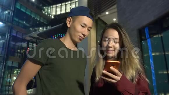 快乐的年轻亚洲夫妇的男朋友和女朋友晚上站在城市街道上使用智能手机视频的预览图