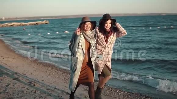 两个女朋友高兴地跑来跑去在露天嬉戏日落时穿着秋装和帽子的年轻女子在海滩上视频的预览图