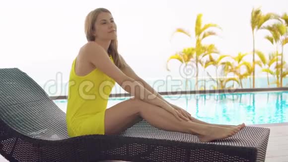 一位穿着黄色衣服躺在绿松石水游泳池旁的日光浴池里的美丽梦幻的年轻女子视频的预览图