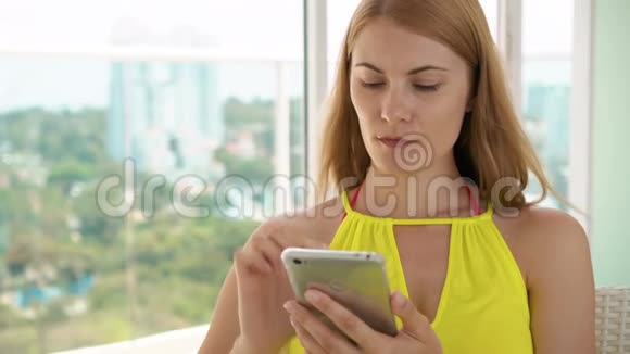 微笑快乐的女人穿着黄色的裙子坐在阳台上用她的智能手机和朋友聊天视频的预览图