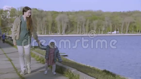 城市公园里的母亲和蹒跚学步的女孩他们牵着手走在湖堤上视频的预览图