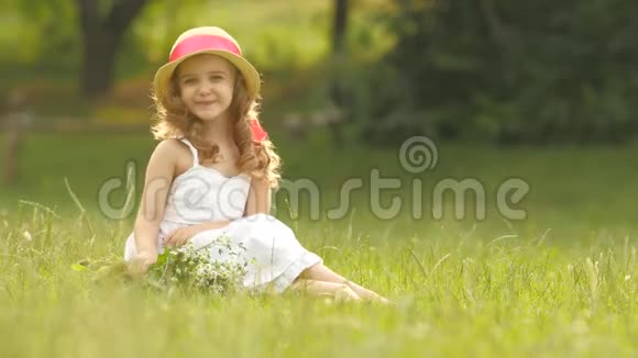 婴儿坐在草地上戴着一顶撒拉芬和帽子手持野花微笑着视频的预览图
