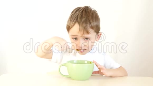 这个孩子很高兴地用勺子吃盘子里的汤一个男孩坐在白色背景的桌子旁吃饭视频的预览图