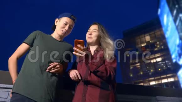 快乐的年轻亚洲夫妇的男朋友和女朋友晚上站在城市街道上使用智能手机视频的预览图