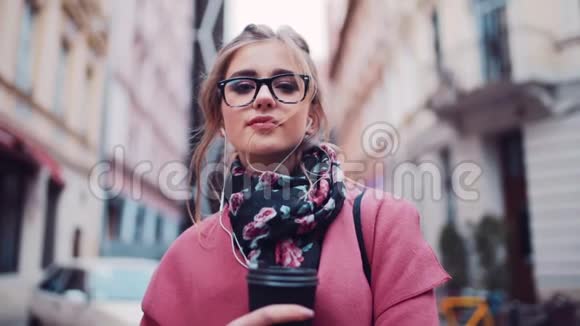 迷人的年轻女子站在城市街道上端着一杯咖啡用耳机听音乐看着视频的预览图