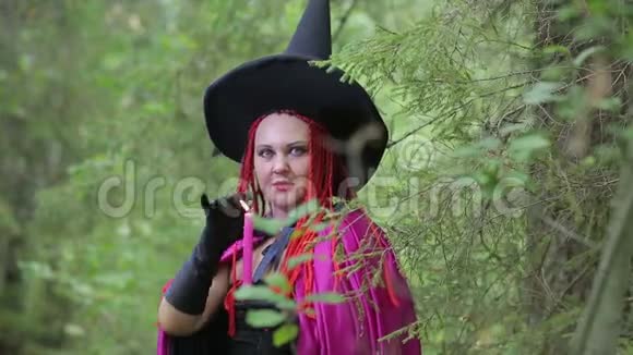 一个年轻的女巫一头红头发一顶尖尖的帽子和一件黑色的斗篷在森林里点燃蜡烛视频的预览图