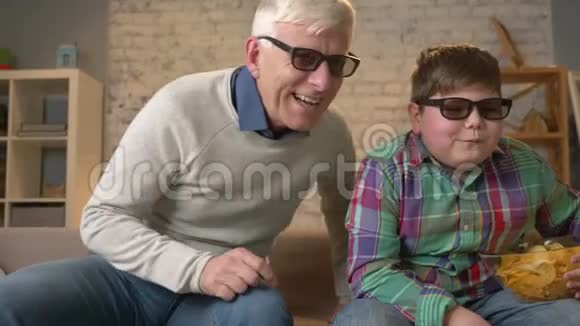 祖父和孙子坐在沙发上用3D眼镜看3D电影吃薯片看电视看节目家庭视频的预览图