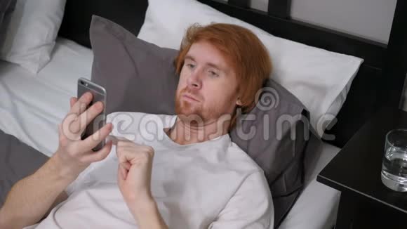 使用智能手机上网躺在床上的红发男子视频的预览图