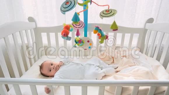 可爱的3个月大的婴儿躺在大窗户旁边的白色木制婴儿床上视频的预览图