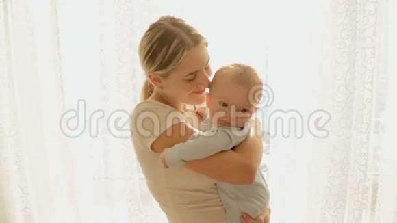 在明亮的阳光下幸福的年轻母亲在大窗前抱着她3个月大的小男孩轻轻地拍了一张温暖的照片视频的预览图