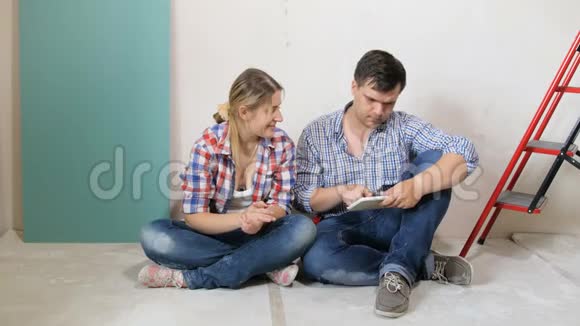 一对年轻夫妇坐在正在装修的公寓地板上在平板电脑上选择家具的4K镜头人与人视频的预览图