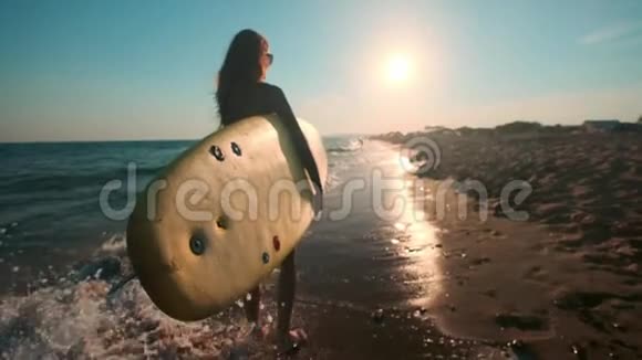 慢动作美丽苗条的女孩走在海滩上拿着冲浪板傍晚日落时跑步害怕视频的预览图