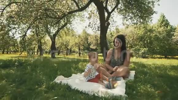 快乐的年轻妈妈和她可爱的儿子在公园的树下玩毯子视频的预览图