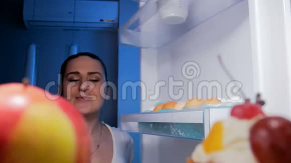 4K镜头年轻微笑的女人在夜里看着冰箱里咬着红苹果视频的预览图