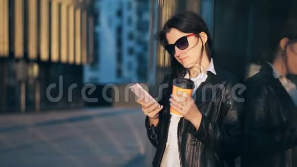 年轻漂亮的女商人带着一杯咖啡正在城市里用智能手机办公大楼视频的预览图