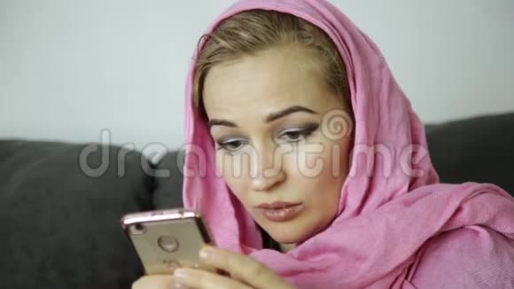 穿着粉红色头巾的年轻穆斯林妇女坐在咖啡馆的沙发上用手机发短信视频的预览图