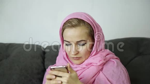 漂亮的阿拉伯女人戴着粉红色头巾坐在咖啡馆的沙发上用手机发短信视频的预览图