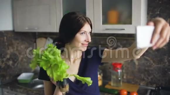 有趣的女人家庭主妇在家里的厨房做饭时用绿色沙拉拍摄自拍视频的预览图