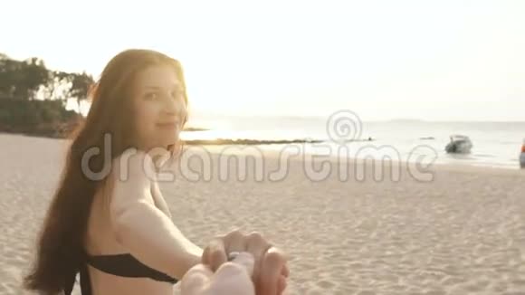 慢动作跟着我拍摄一个穿比基尼的年轻女孩在日落时在海滩上奔跑牵着男人的手视频的预览图